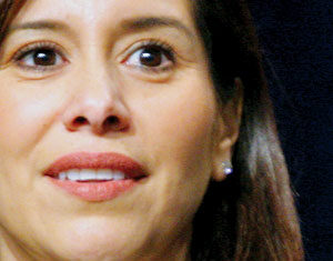A close-up of a woman with long brown hair, wearing a white collared shirt, dark blazer, pearl necklace, and stud earrings, with a neutral expression.