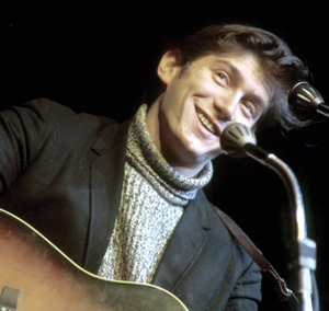 A smiling man in a turtleneck sweater and jacket plays an acoustic guitar and sings into a microphone on stage.