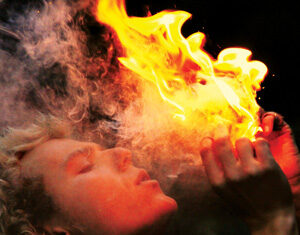 A person with curly blond hair exhales smoke or breathes out while holding a bright, orange flame close to their mouth against a dark background.