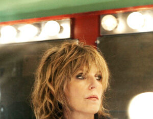 A woman with tousled, shoulder-length blonde hair and dark eye makeup stands in front of a mirror framed with round lights, wearing layered necklaces and a black jacket.