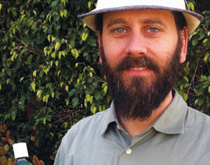 A bearded man in a light-colored hat and shirt stands outdoors, holding a small bottle with a label in his right hand. Green foliage fills the background.