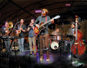 A seven-member band performs on stage with various instruments, including guitar, trumpet, saxophone, trombone, keyboard, drums, and upright bass, under a curved canopy with stage lights overhead.