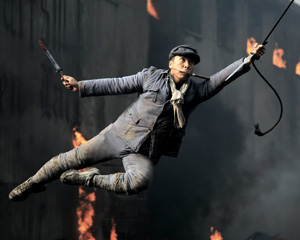 A person wearing a cap and scarf swings through the air holding a sword and a rope, surrounded by flames and a smoky, burning building in the background.