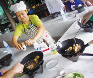 A woman wearing a chef’s hat and apron is cooking with a wok, seasoning food as others cook beside her on a white kitchen counter with cooking utensils and ingredients visible.