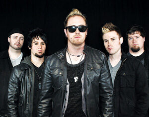 Five men with serious expressions stand together against a black background, all wearing dark jackets. The man in the center wears sunglasses and a necklace, and the group appears to be part of a rock band.