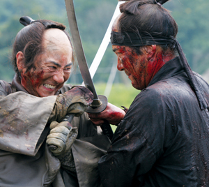 Two men dressed as samurai, faces and clothes smeared with blood, intensely clash swords in close combat, set outdoors with blurred greenery in the background.