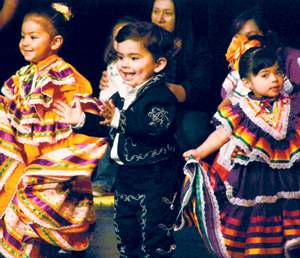 Three young children in colorful traditional Mexican clothing perform a dance. Two girls wear vibrant dresses with ribbons, and a boy wears a black mariachi-style suit. People smile and watch in the background.