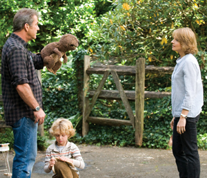 A man holding a beaver puppet faces a woman outdoors, while a young blond child sits on the ground between them. They are surrounded by greenery and a wooden fence.