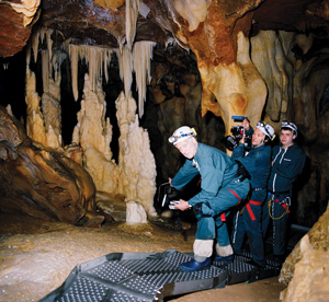 Three people wearing helmets and caving gear film stalagmites and stalactites inside a rocky cave. They stand on a metal walkway, surrounded by dramatic cave formations.