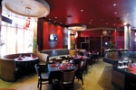Elegant restaurant interior with red walls, round booths, and tables set with red napkins and glassware. Large windows let in natural light, and modern lighting fixtures hang from the ceiling.