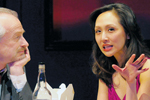 A woman in a pink top gestures with her hand while speaking to a man with grey hair, who is resting his chin on his hand. They are seated at a table with a bottle and a box in front of them.
