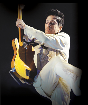 A musician in a white outfit energetically plays an electric guitar, with one leg raised mid-jump against a dark background.