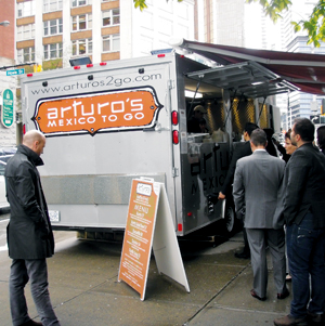 A group of people stand in line at the window of a silver food truck labeled arturos MEXICO TO GO parked on a city street, with a menu board displayed on the sidewalk.
