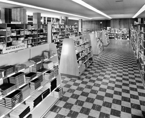Black and white photo of a bookstore or library with checkered floor, shelves lined with books, and a sign labeled Mathematics on one section. The space is well-lit and organized.