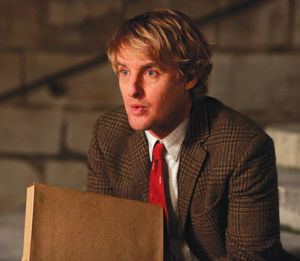 A man with blonde hair wearing a brown plaid jacket, white shirt, and red tie sits holding a brown folder, looking surprised or deep in thought with a stone wall in the background.