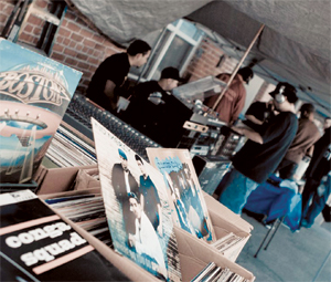 Boxes of vinyl records on a table at an outdoor market, with people browsing and chatting under a canopy in the background. Visible album covers feature various musicians and band names.