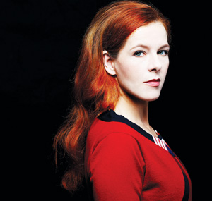 A woman with long red hair and fair skin stands against a black background, looking slightly to the side. She wears a red top and has a calm, confident expression.