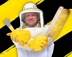 A beekeeper in protective gear and yellow gloves stands with arms crossed, holding a hive tool and a large brush. The background features bold yellow and black diagonal stripes.