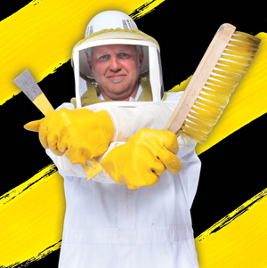 A beekeeper in protective gear and yellow gloves stands with arms crossed, holding a hive tool and a large brush. The background features bold yellow and black diagonal stripes.
