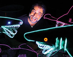 A smiling man is surrounded by several large shark figures outlined in colorful neon lights, creating a glowing effect in the dark.
