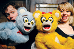 Two people smile and pose with large, colorful bear puppets—one blue with a red bow tie, the other yellow with a red hair bow. Both puppets have exaggerated facial expressions.