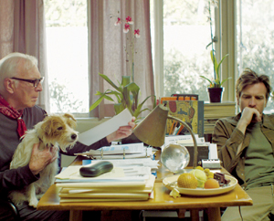An older man holding a small dog sits at a cluttered desk with papers and fruit, while a younger man sits nearby, looking thoughtful. Large windows and plants are in the background, letting in soft natural light.