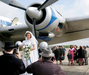 A bride in a white gown and veil poses with a bouquet in front of a vintage airplane as photographers take her picture. A group of people, some with luggage, stand in the background near the plane.