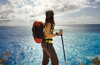 A person with a backpack and hiking pole stands on a cliff, looking out over a bright blue ocean under a partly cloudy sky.