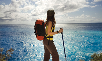 A person with a backpack and hiking pole stands on a cliff, looking out over a bright blue ocean under a partly cloudy sky.