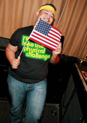 A smiling person wearing a yellow headband and a black T-shirt that says Ill take the physical challenge holds an American flag indoors, standing in front of tan curtains and audio equipment.
