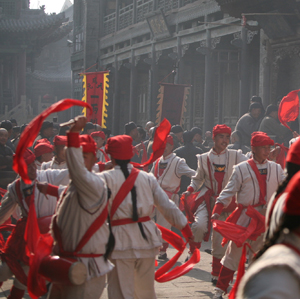 A group of performers in white costumes with red sashes and hats dance energetically in a street, waving red ribbons. Banners and a crowd of onlookers are visible in the background, set in a traditional-looking area.