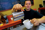 A barista wearing a black shirt pours steamed milk from a metal pitcher into a white mug, standing behind a counter with a chalkboard menu in the background.
