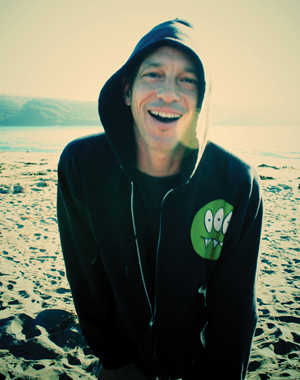 A person wearing a black hoodie with a green cartoon face stands smiling on a sandy beach, with sunlight shining and water and hills in the background.