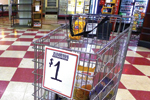A metal bin inside a store displays various packaged goods for sale, with a large sign reading BARGAINS $1 attached to the front. The store has red and white checkered flooring.