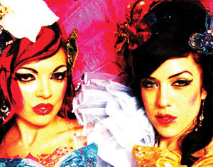 Two women with dramatic makeup and colorful costumes pose in front of a bright pink background. Each holds a large butterfly decoration, one blue and one yellow, and they wear elaborate accessories and ruffled details.