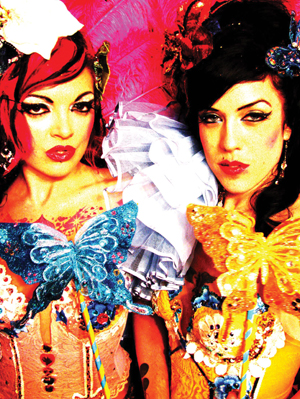Two women with dramatic makeup and colorful costumes pose in front of a bright pink background. Each holds a large butterfly decoration, one blue and one yellow, and they wear elaborate accessories and ruffled details.