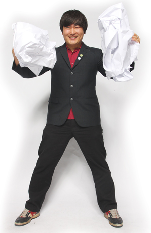 A person in a black suit and red shirt stands smiling, holding large crumpled sheets of white paper in both hands, with legs spread apart against a white background.