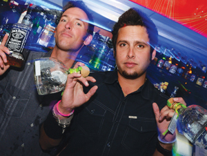 Two men in a bar hold bottles of liquor with colorful lights in the background, appearing to enjoy a lively night out. Shelves filled with bottles are visible behind them.