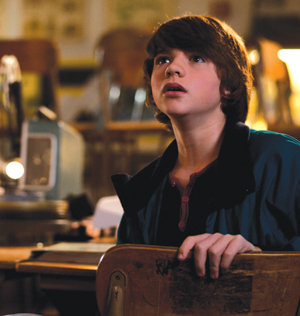 A young boy with brown hair sits in a classroom, looking upward with a surprised or curious expression. He is holding the back of a wooden chair, and old classroom equipment is visible in the background.