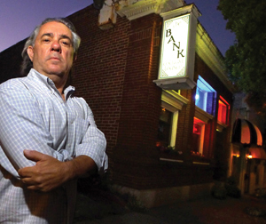 A man with gray hair and a checked shirt stands with his arms crossed in front of a brick building with a lit sign that reads BANK and colorful lights in the windows.