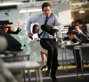 A man in office attire runs frantically through an office as glass shatters and debris flies, while other people duck and shield themselves at their desks.
