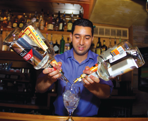 A bartender in a blue shirt pours liquor from two bottles into a glass filled with ice at a bar with shelves of bottles in the background.