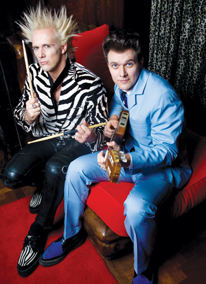 Two men sit on a red couch; one with spiky blond hair, wearing a zebra-striped jacket and holding drumsticks, and the other with dark hair in a light blue suit, holding a guitar.