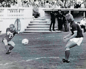 Black and white photo of a soccer match; one player is kicking the ball while another faces him. Spectators and officials watch from the stands in the background.