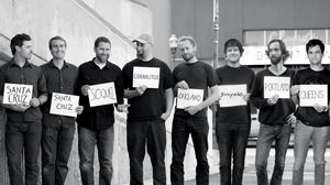 Eight men stand in a line outdoors, each holding a sign with the name of a different city: Santa Cruz, Soquel, Corralitos, Oakland, Sunnyvale, Portola Valley, and Queens. The photo is black and white.
