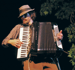 A person wearing a hat and sunglasses plays an accordion outdoors at night, with tree branches visible in the background.