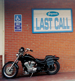 A black motorcycle is parked on a sidewalk in front of a brick wall with a sign that says LAST CALL. A blue handicapped parking sign is mounted on the wall above the motorcycle.