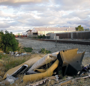 A pile of discarded furniture and debris lies beside a railroad track, with industrial buildings and a cloudy sky in the background.