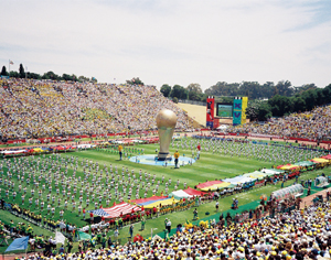 A crowded stadium hosts a colorful opening ceremony with performers surrounding a large World Cup trophy replica at the center of the field, and spectators filling the stands.