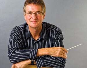 A man with glasses wearing a black and white striped shirt sits on a chair backwards, smiling and holding a conductors baton in his right hand against a plain gray background.
